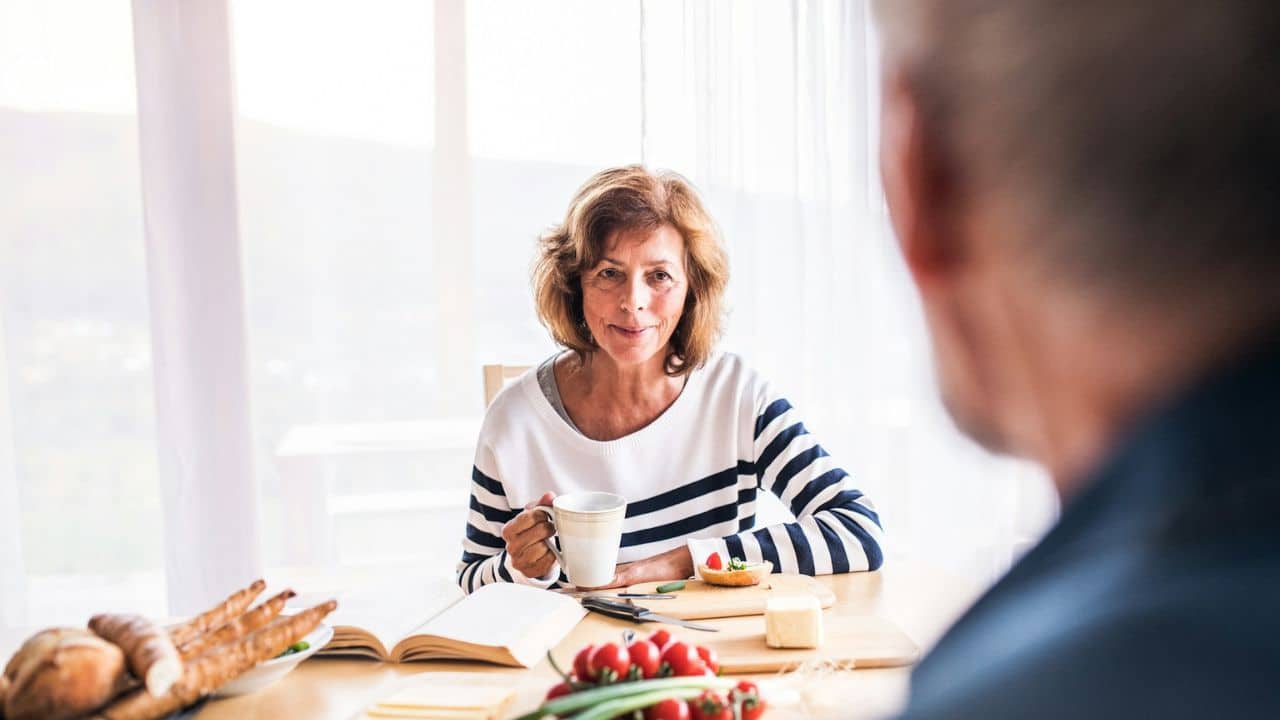 A woman with curly brown hair holds a mug and looks at someone across a table.