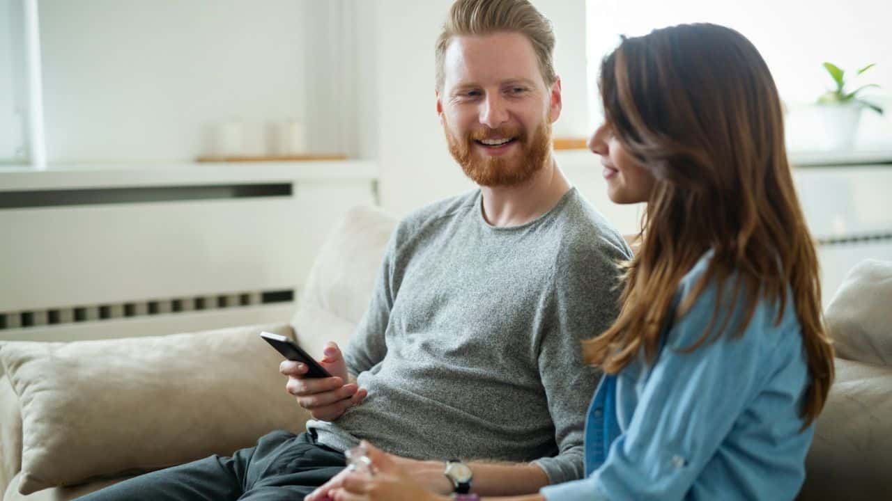A man with a red beard smiles and holds a phone while looking at a woman on a couch.