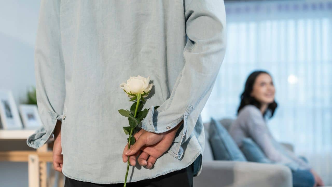 A person holds a white rose behind their back, facing away from a woman sitting on a couch.
