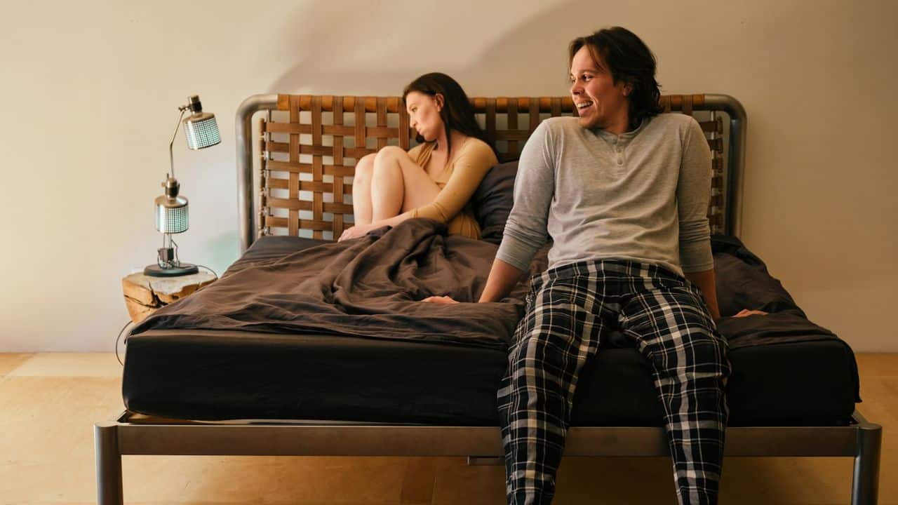 A man in plaid pajama pants sits on a bed, looking to the right, while a woman sits further back, looking away.