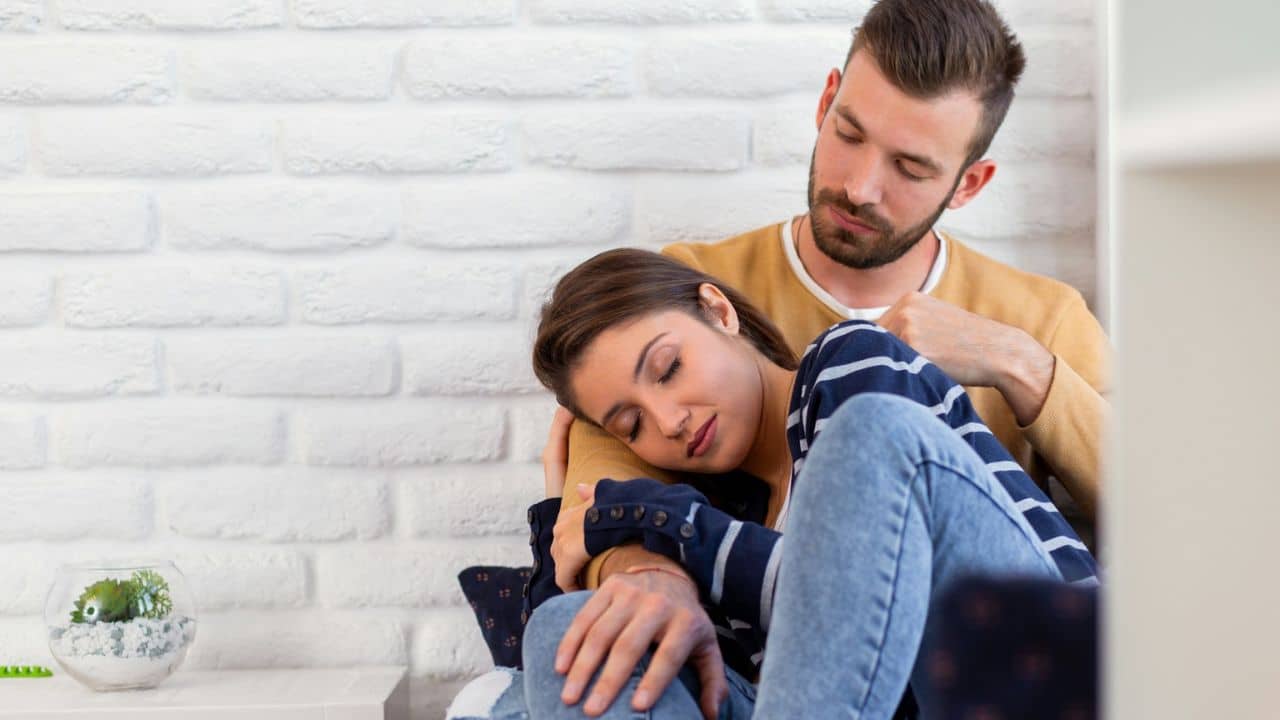 A man with a beard sits behind a woman, who is resting her head on his arm.