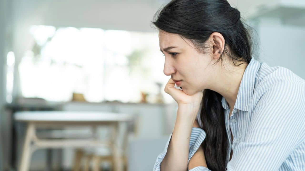 A young woman with long dark hair holds her chin, looking down with a worried expression.