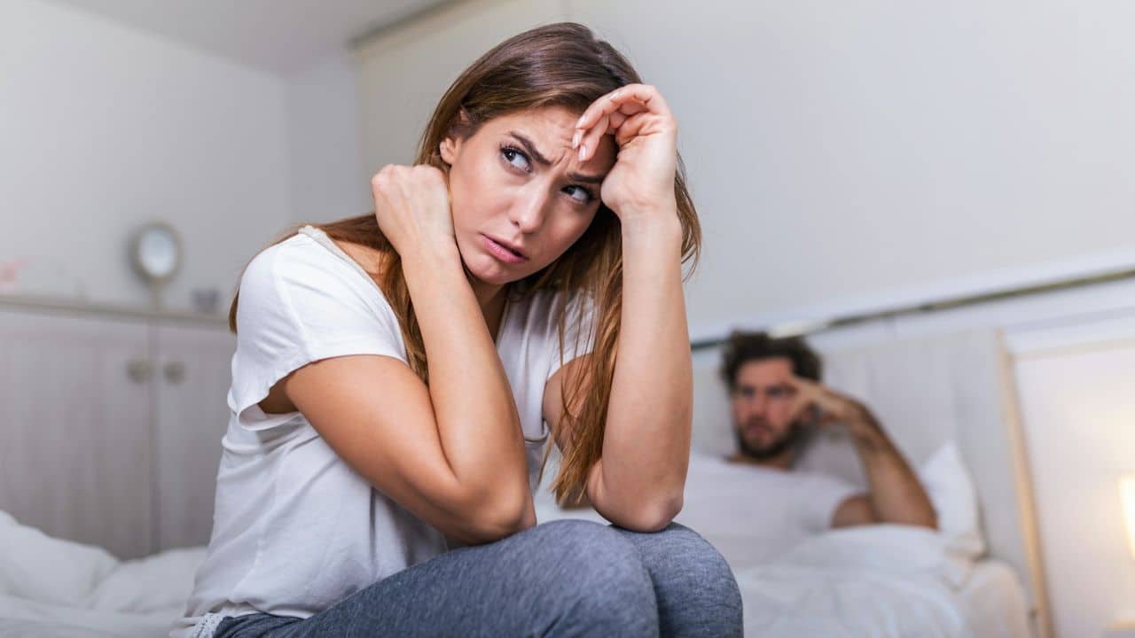 A woman sits on a bed with a worried expression, while a man is out of focus in the background.