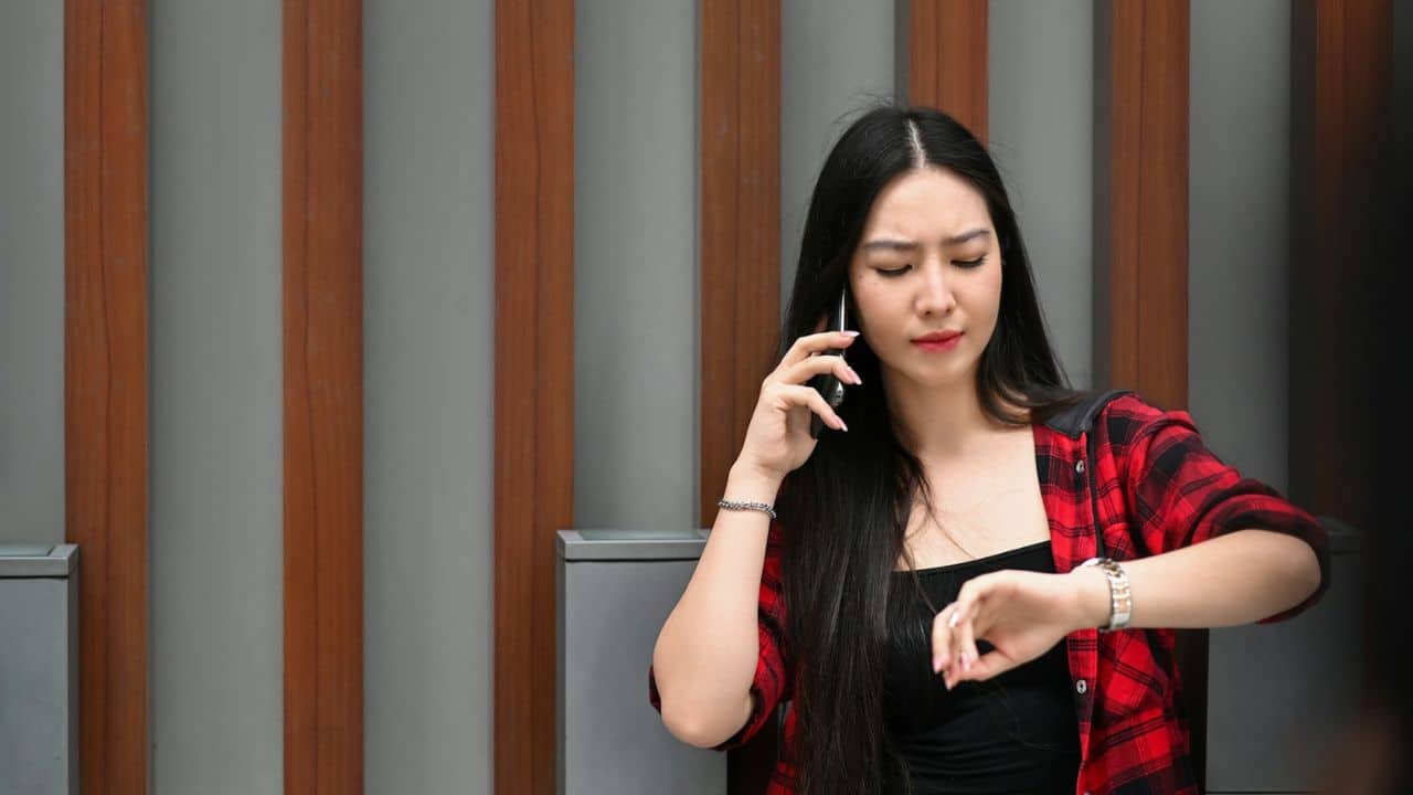 A woman with long black hair talks on a phone and checks her watch.