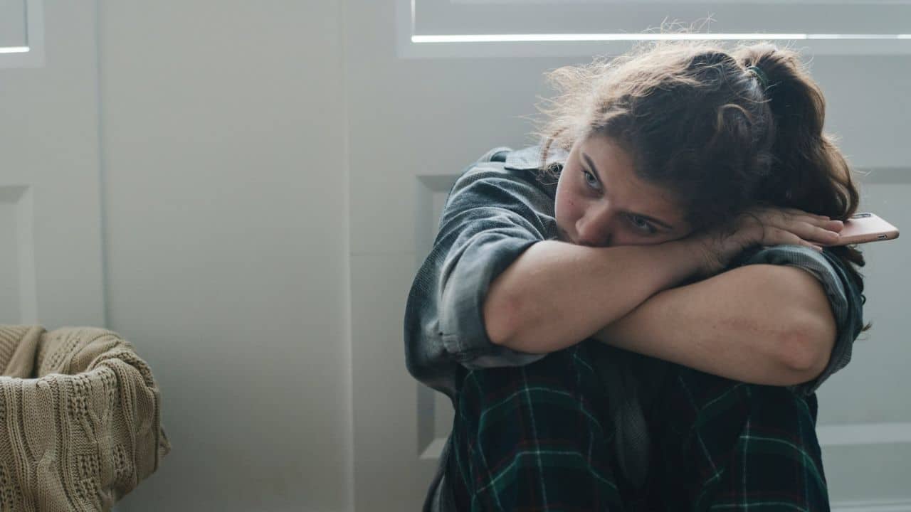 A person with brown hair in a ponytail sits on the floor, hugging their knees, looking sad.