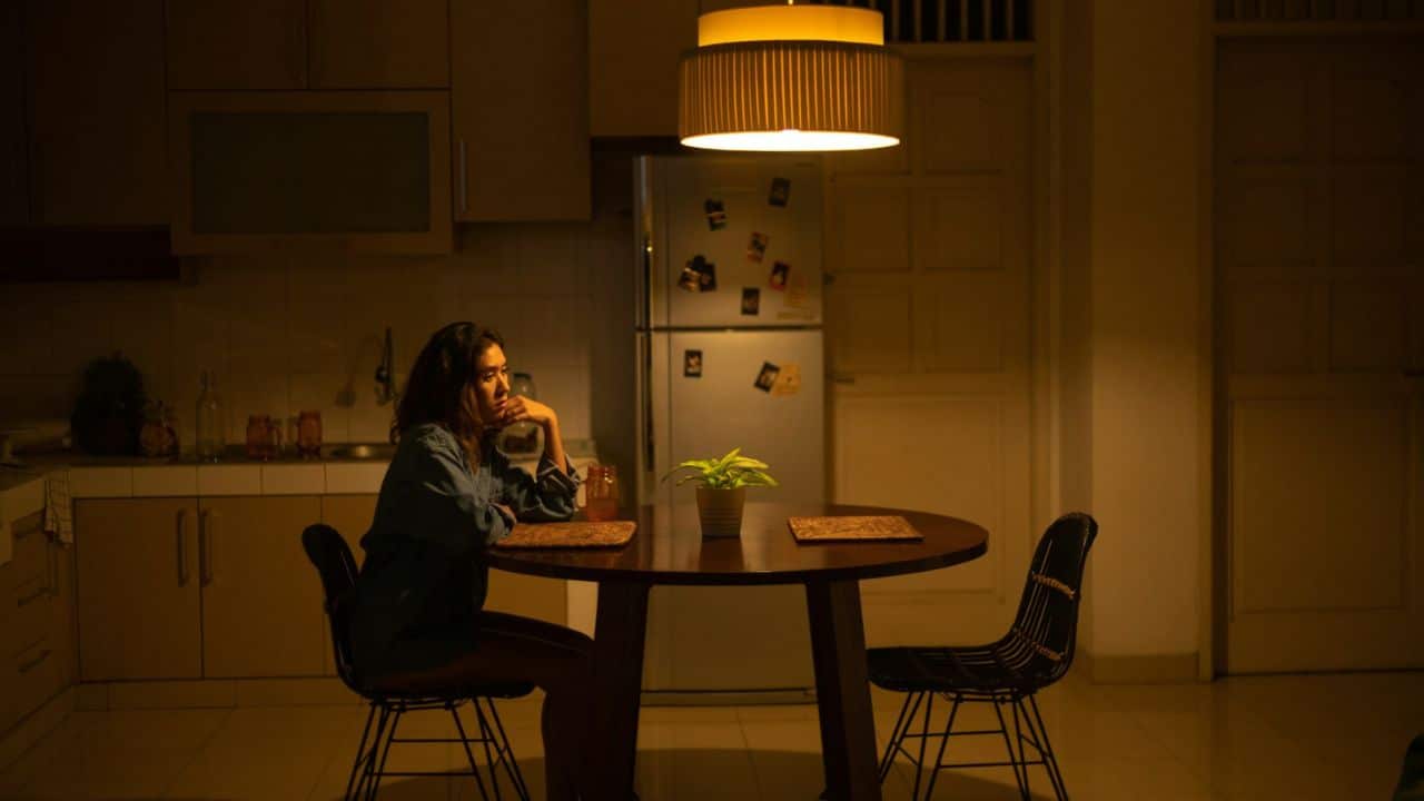 A woman sits alone at a dining table in a dimly lit kitchen.