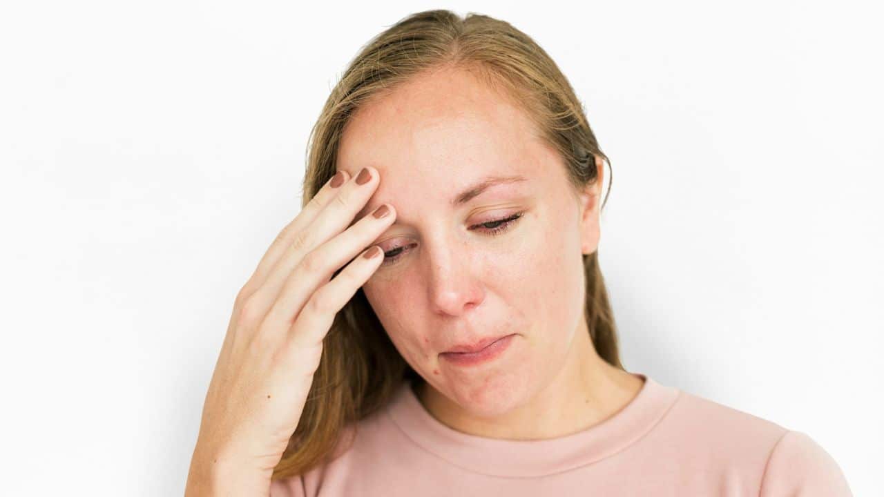 A woman with light brown hair looks down, touching her forehead with her hand.