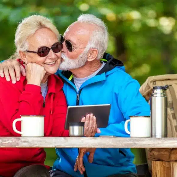 An elderly man in sunglasses kisses a smiling elderly woman on the cheek, holding a tablet.