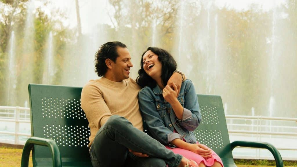 A man and woman laugh together on a green park bench, with a fountain in the background.