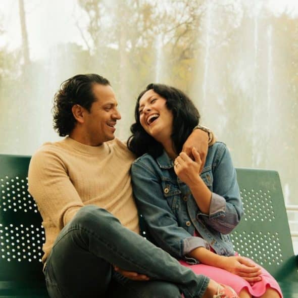 A man and woman laugh together on a green park bench, with a fountain in the background.
