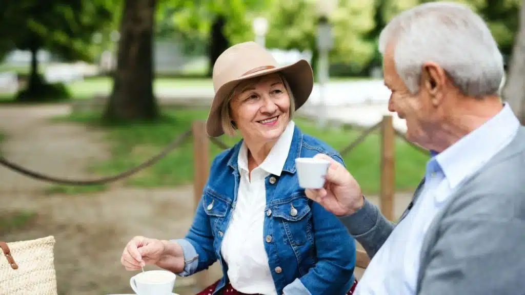 An elderly woman in a hat and denim jacket smiles at an older man while holding a cup.