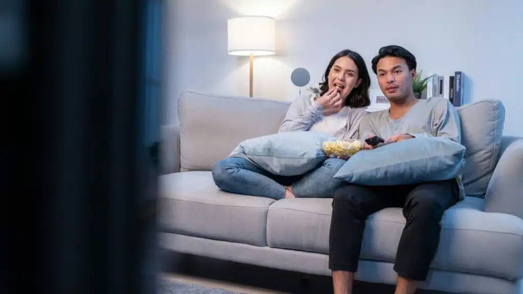 A man and a woman sit on a couch, watching TV and eating popcorn.