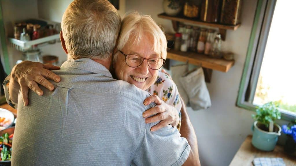 A smiling elderly woman with glasses hugs a man in a kitchen.