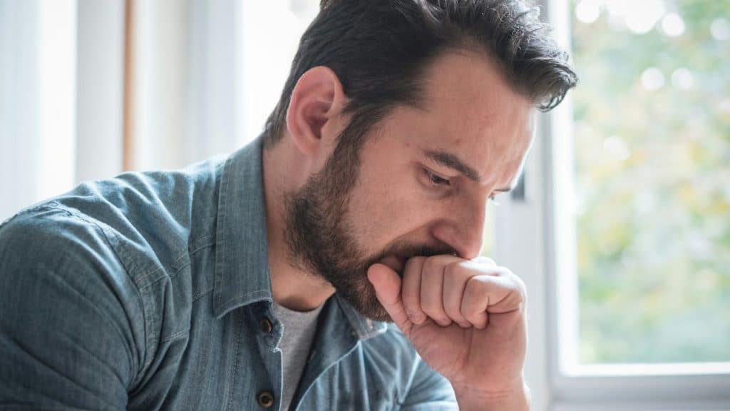 A bearded man in a denim shirt looks down, deep in thought, with his hand to his mouth.