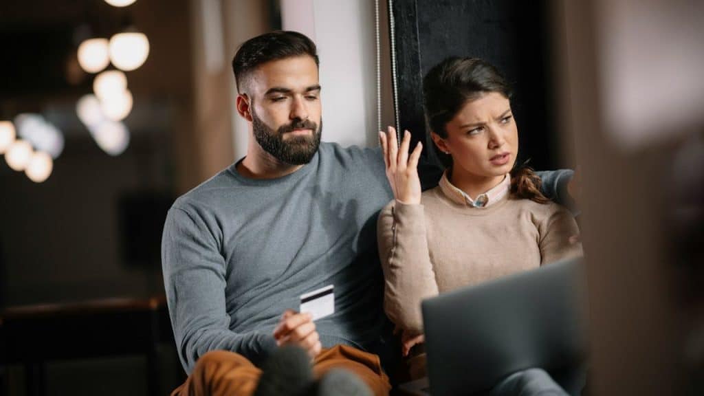 A man with a beard holds a credit card while looking at a woman with a laptop.