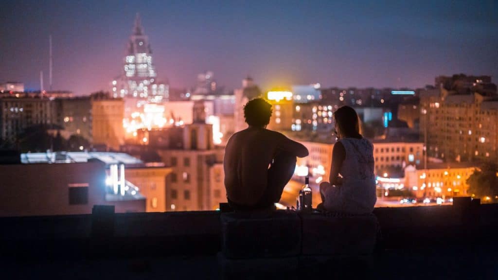 A shirtless person and another person sit on a rooftop, overlooking a city skyline at night.