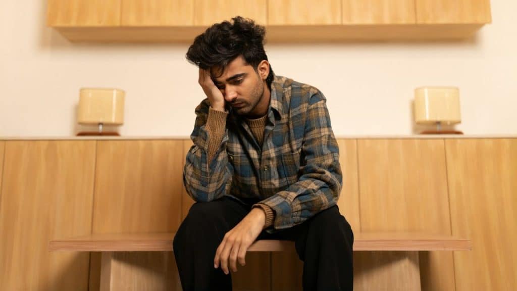 A man in a plaid shirt sits, looking stressed with his hand on his head.