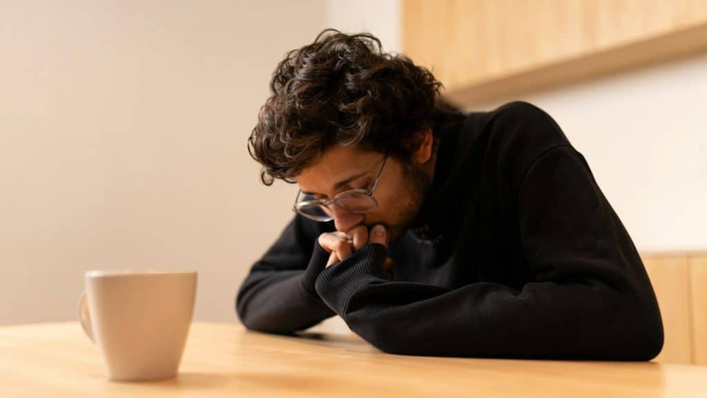 A man with curly hair and glasses sits at a table, hands clasped, looking down.