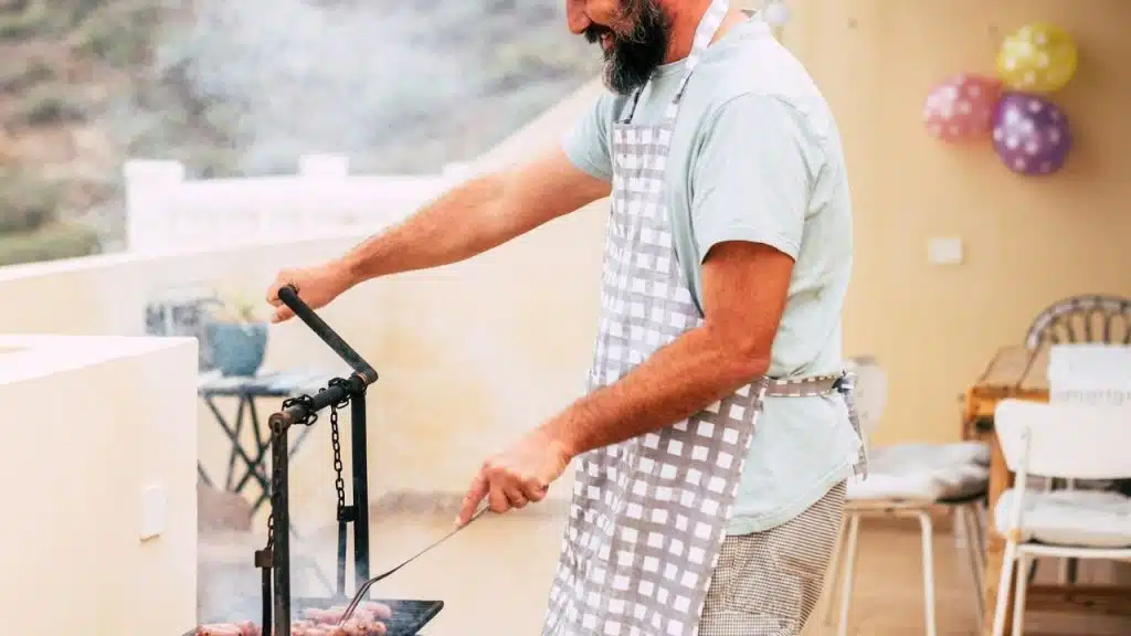 A man in an apron is grilling meat outdoors, with smoke rising from the grill.
