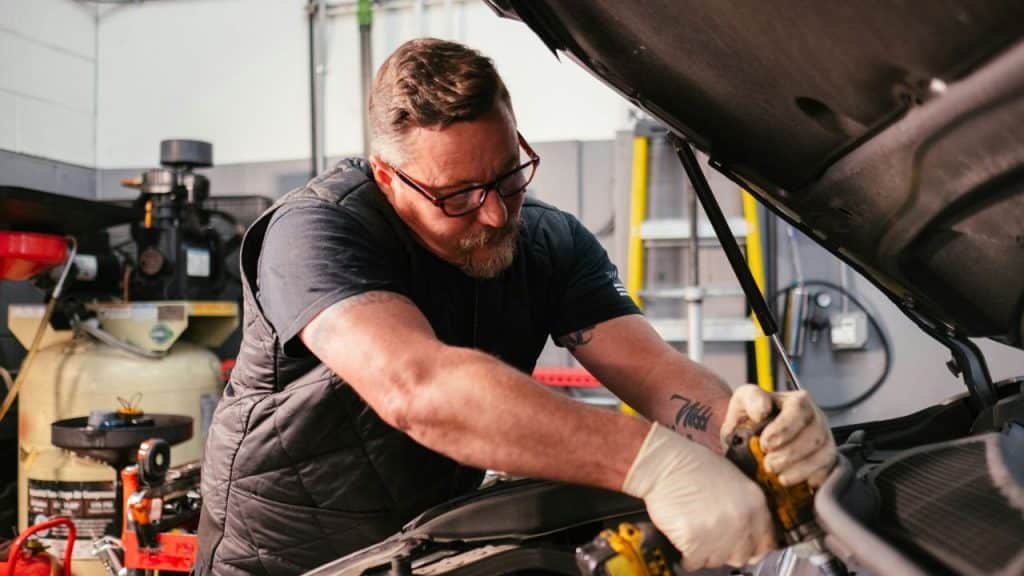 A bearded man wearing glasses and gloves works on an open car engine in a garage.