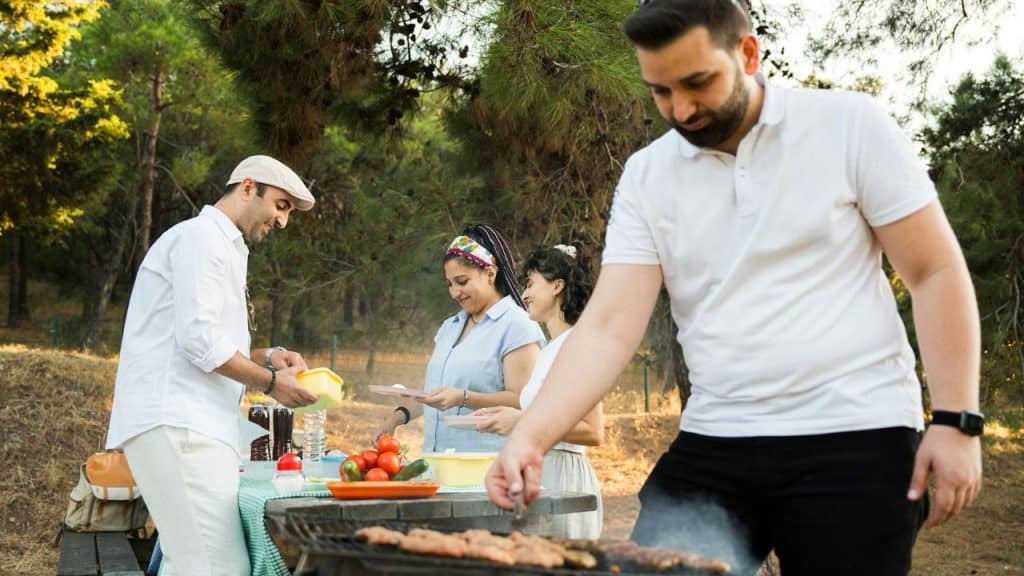 A group of friends gathers around a grill outdoors, preparing food with a man grilling in the foreground.