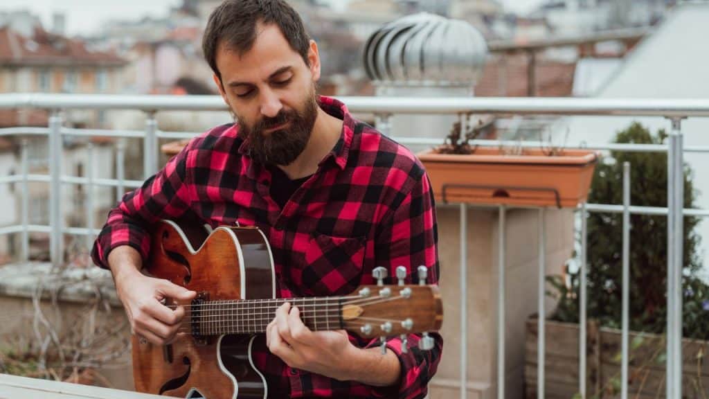 A bearded man in a red plaid shirt plays an acoustic guitar on a balcony.