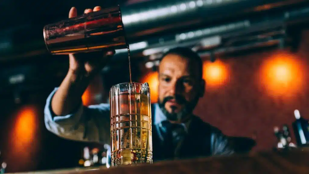A bartender with a beard pours a drink from a shaker into a tall glass with ice.