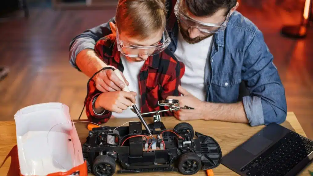 A father and son, wearing safety goggles, work together soldering components on a model car.