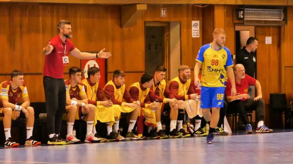 A coach in a red shirt gestures to a player walking past the bench during a game.