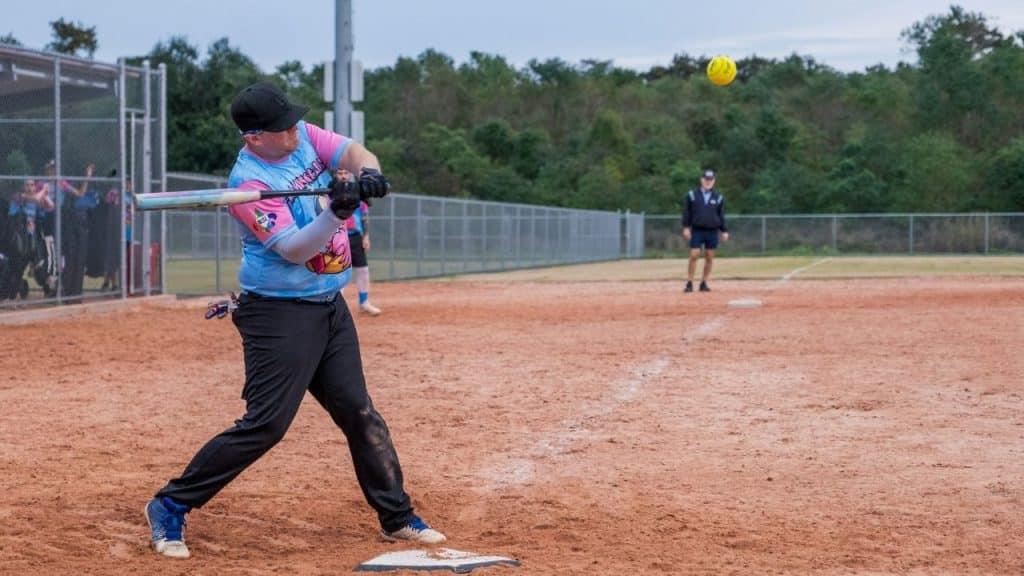 A softball player in a blue and pink jersey swings a bat, hitting a yellow ball.