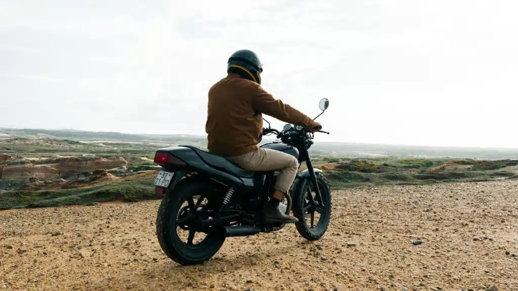 A motorcyclist wearing a helmet and brown jacket sits on a motorcycle on a dirt road, overlooking a landscape.