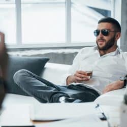 A bearded man in sunglasses lounging on a sofa with a glass of whiskey.
