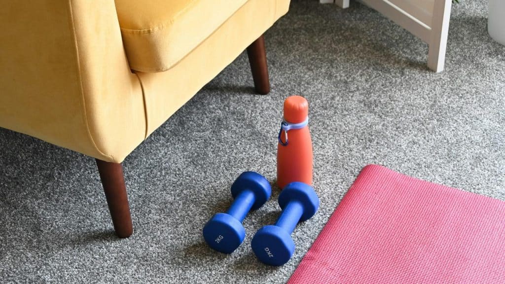 A pair of blue dumbbells and an orange water bottle beside a red exercise mat.
