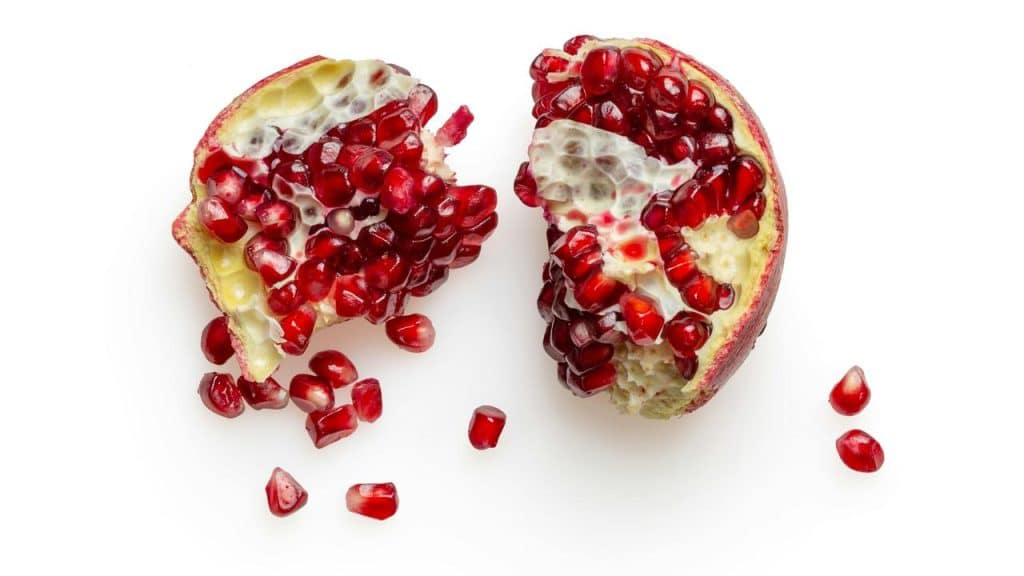 A halved pomegranate with glossy red seeds spilling onto a white surface.