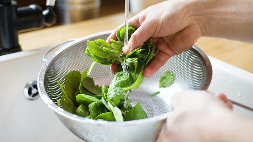 A person rinsing fresh spinach leaves in a metal colander under running water.