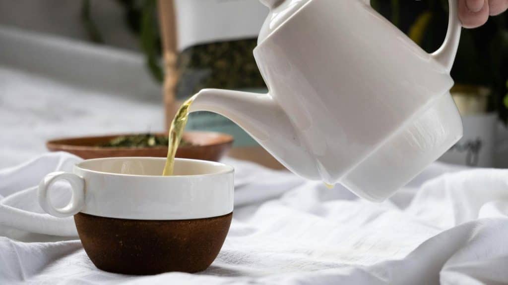 A white teapot pouring hot tea into a matching cup on a linen-covered table.