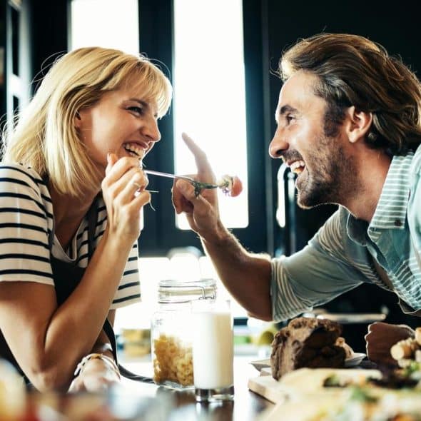 An affectionate couple laughing and feeding each other while cooking in a cozy kitchen.