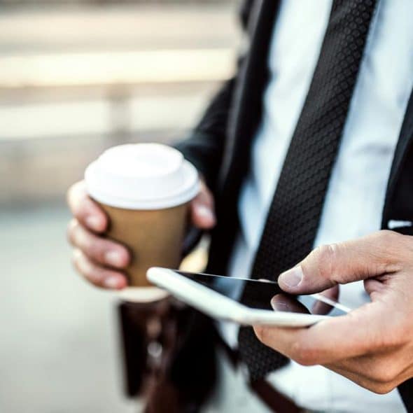 A person in a suit holding a takeaway coffee cup and tapping on a smartphone.
