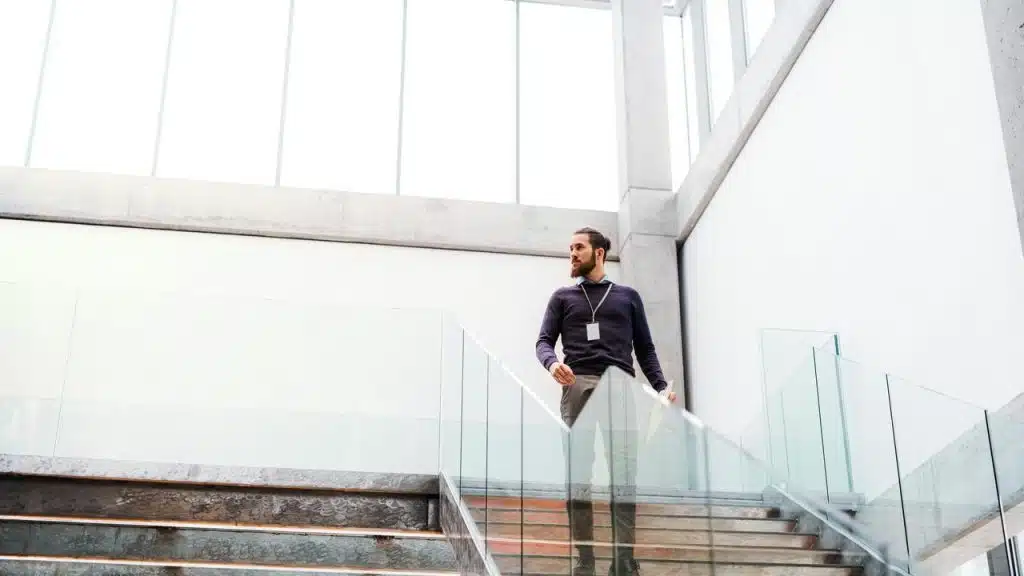 A bearded man with a lanyard walking down a glass-and-concrete staircase in a bright atrium.
