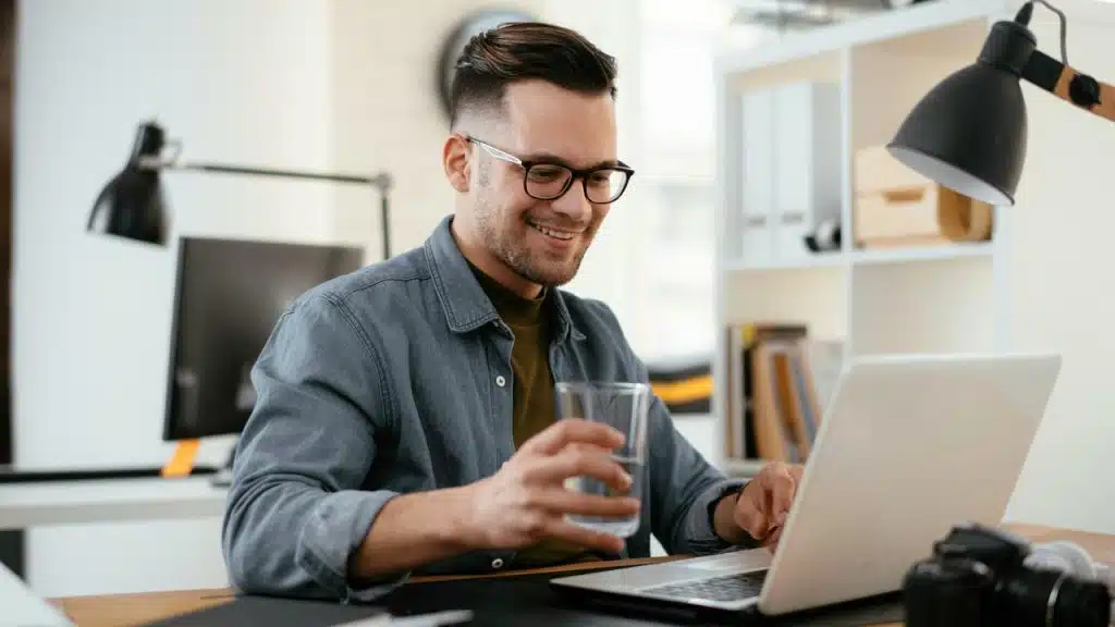 A smiling man wearing glasses holding a glass of water while typing on a laptop at his desk.