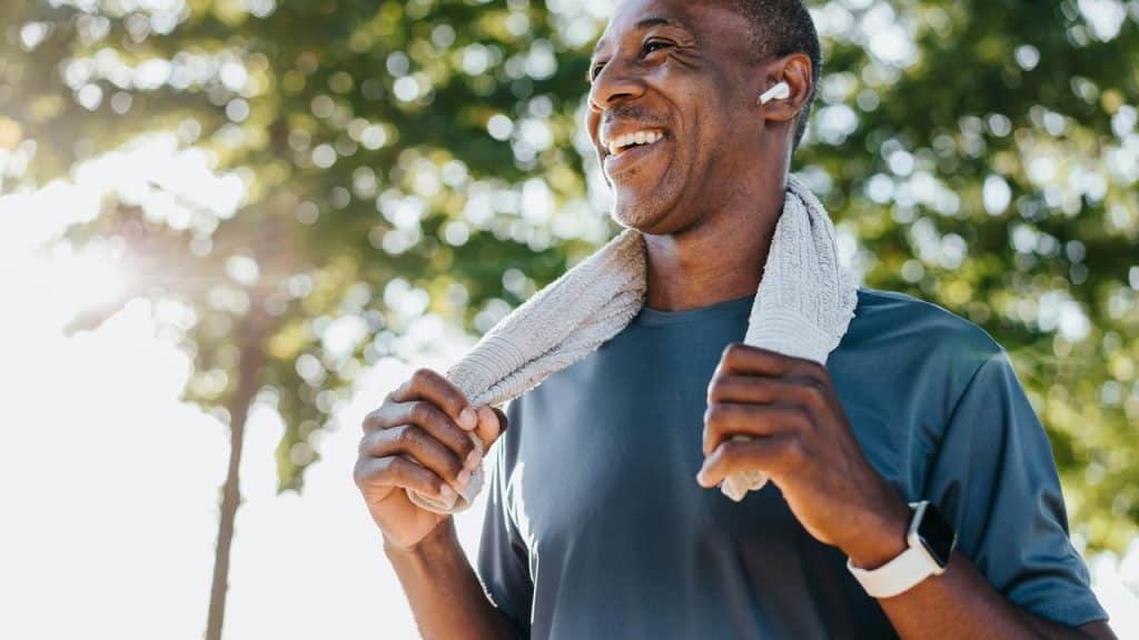 A smiling man in athletic gear wearing earbuds with a towel draped around his shoulders.