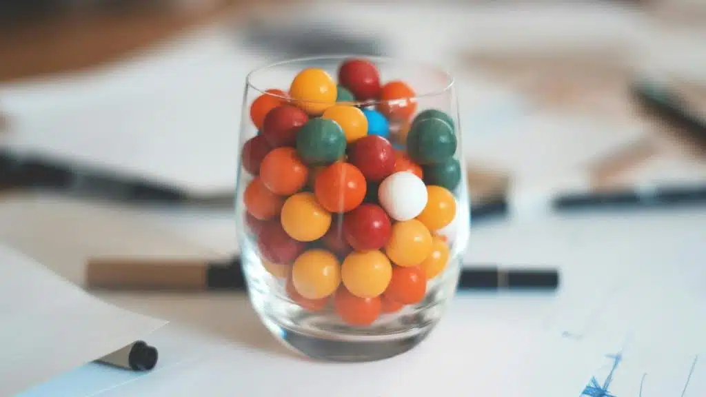 A clear glass filled with multicolored chewing gum sitting on a desk.