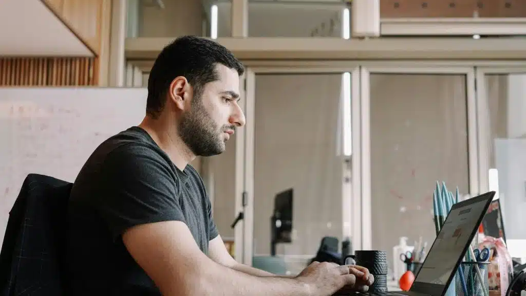 A bearded man typing on a laptop at a cluttered office desk.