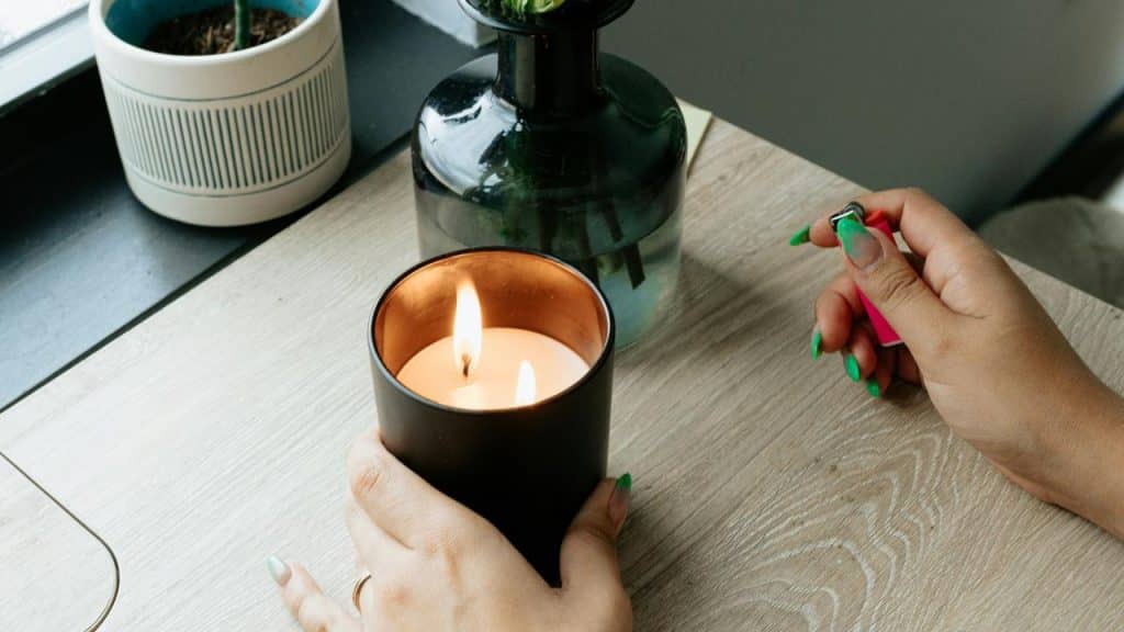 A person’s hands lighting a two-wick candle in a black glass jar.