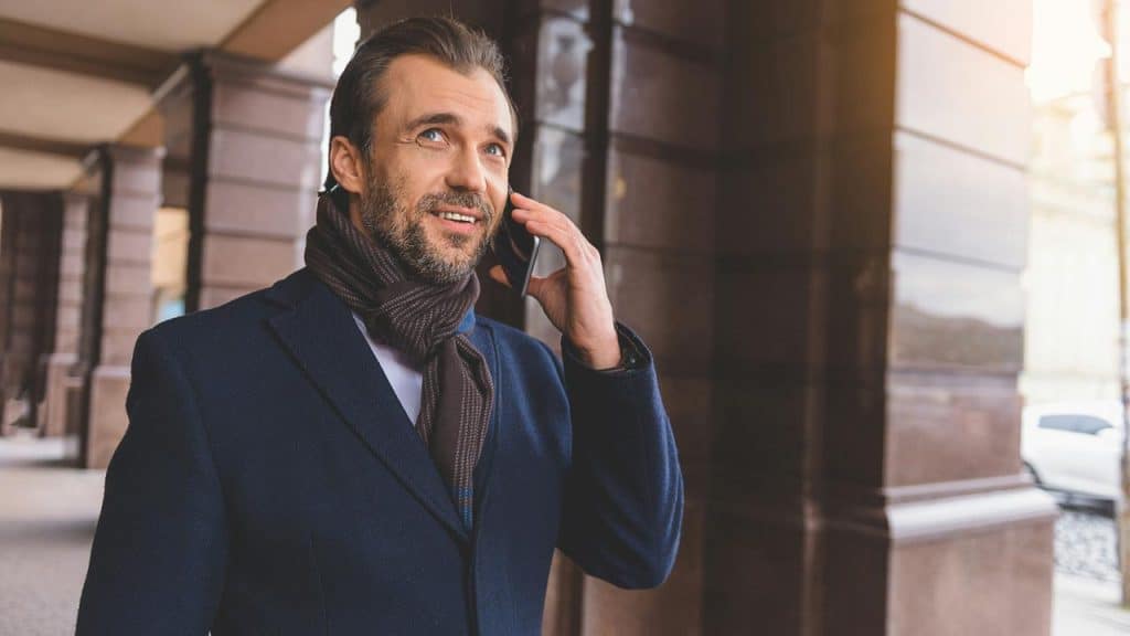 A bearded man in a coat and scarf talking on a smartphone under a colonnade.
