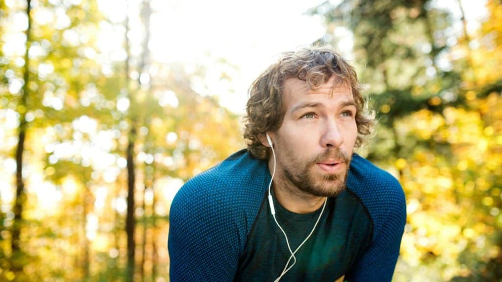 A man with earbuds catching his breath in a sunlit forest.