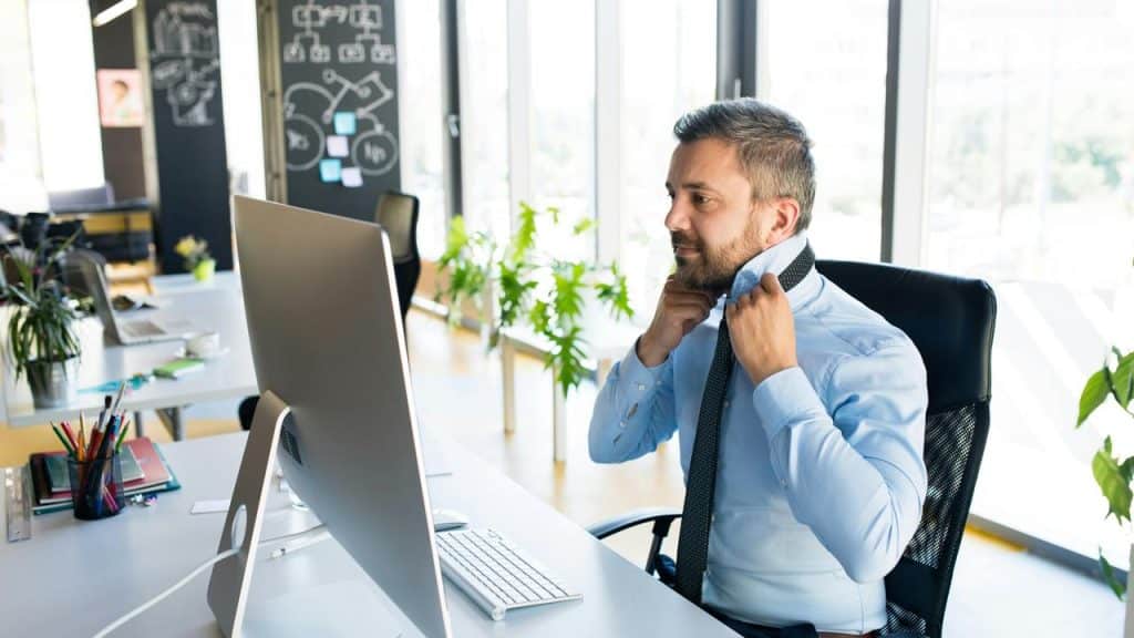 A man at an office desk ties his necktie in front of a computer.
