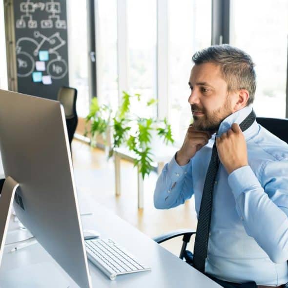 A man at an office desk ties his necktie in front of a computer.