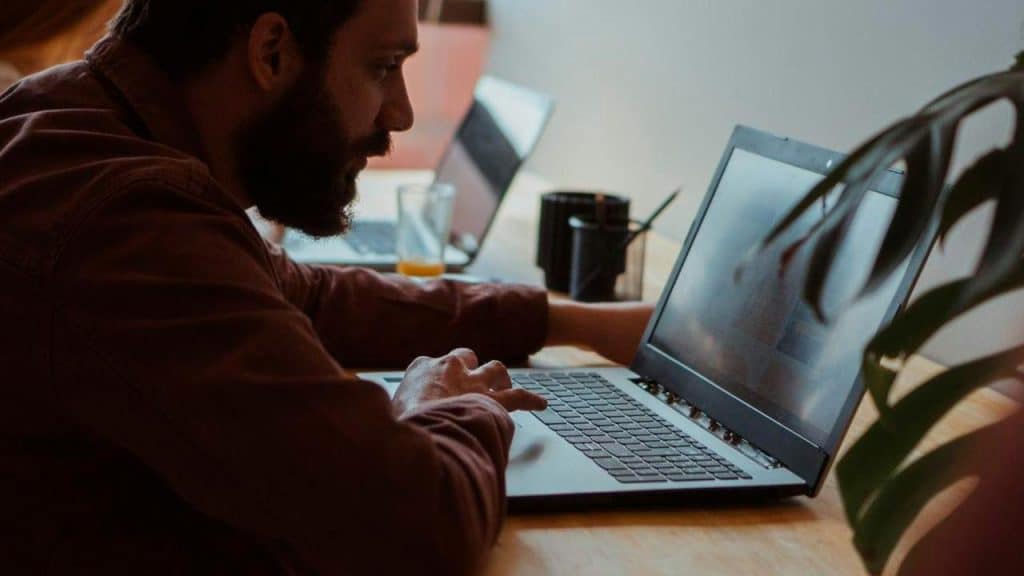 A bearded man typing on a laptop at a wooden desk.