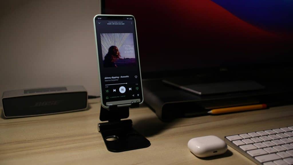 A smartphone on a desk stand playing music beside a keyboard and speaker.